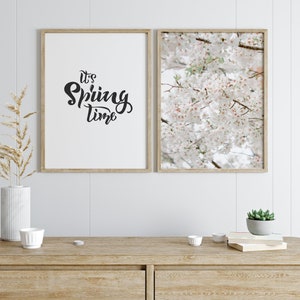 May include: Two framed prints on a white wall. The print on the left says "It's Spring Time" in black script. The print on the right shows a close-up of white cherry blossoms.