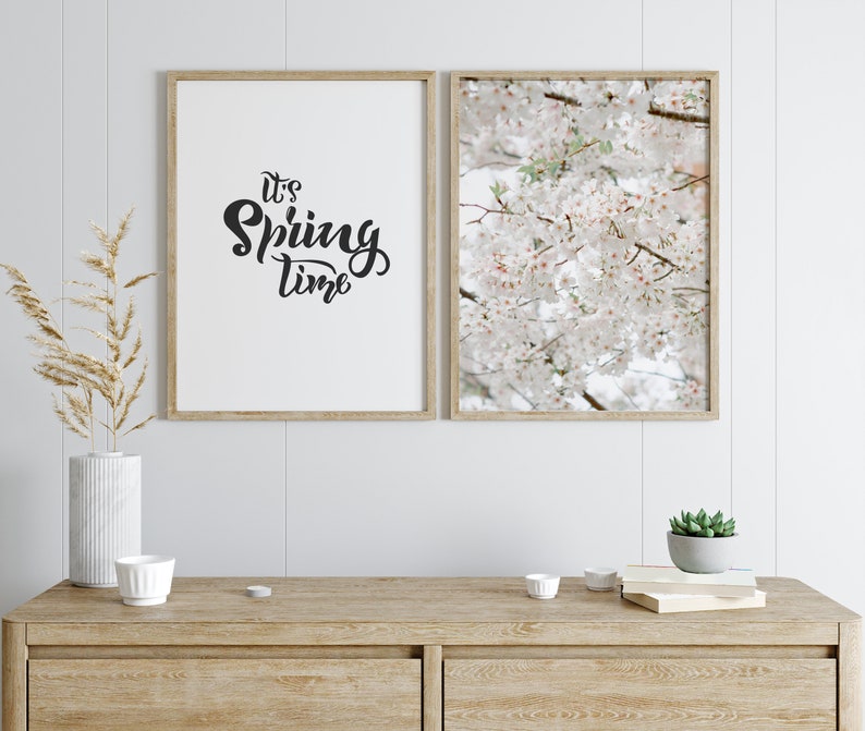 May include: Two framed prints on a white wall. The print on the left says "It's Spring Time" in black script. The print on the right shows a close-up of white cherry blossoms.