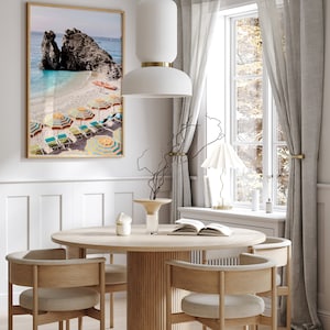 Puede incluir: Una mesa de comedor redonda de madera con una base acanalada y cuatro sillas. Las sillas tienen marcos de madera marr&oacute;n claro y cojines beige. Un jarr&oacute;n de cer&aacute;mica blanco con una rama seca se encuentra sobre la mesa, junto con un libro y una vela blanca. Una impresi&oacute;n enmarcada de una escena de playa con sombrillas coloridas cuelga en la pared detr&aacute;s de la mesa.
