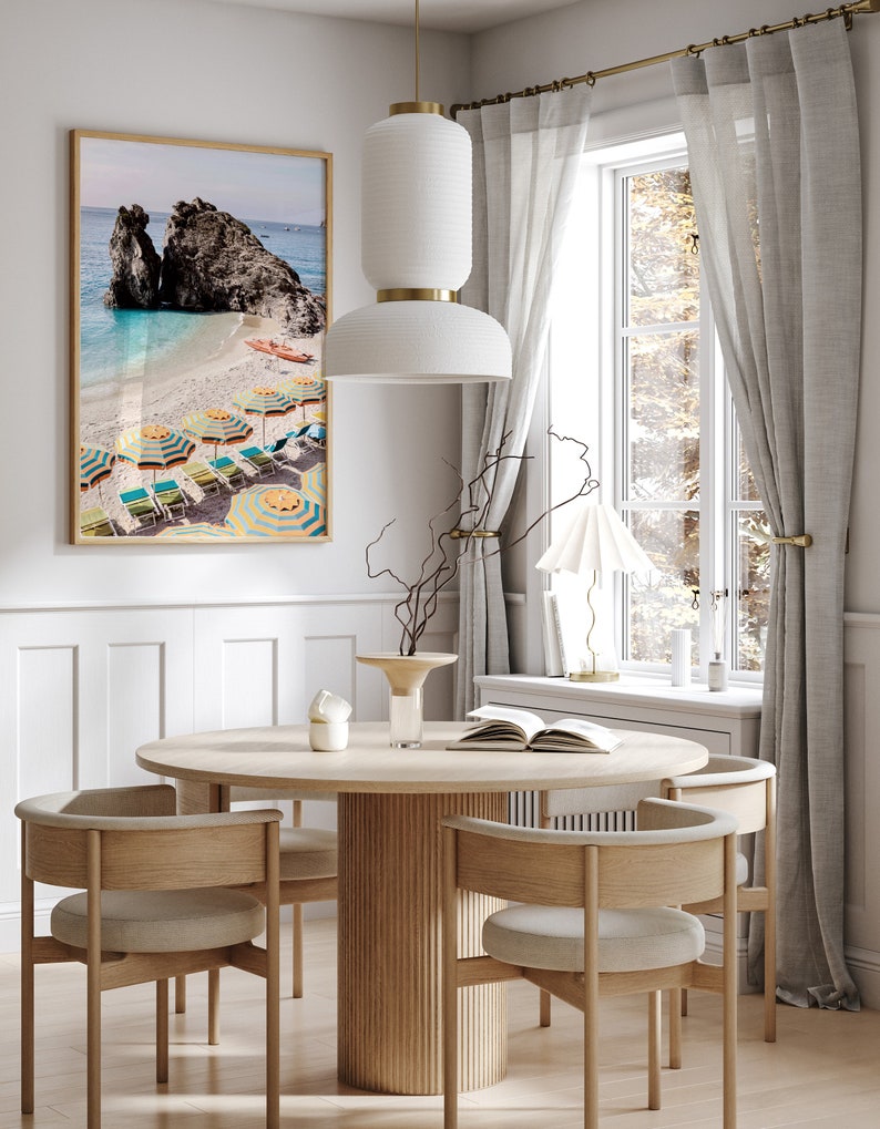 Puede incluir: Una mesa de comedor redonda de madera con una base acanalada y cuatro sillas. Las sillas tienen marcos de madera marr&oacute;n claro y cojines beige. Un jarr&oacute;n de cer&aacute;mica blanco con una rama seca se encuentra sobre la mesa, junto con un libro y una vela blanca. Una impresi&oacute;n enmarcada de una escena de playa con sombrillas coloridas cuelga en la pared detr&aacute;s de la mesa.