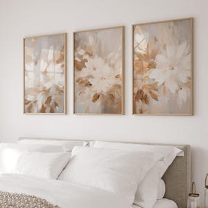 May include: Three framed abstract art prints with a beige background and white floral designs. The prints are arranged in a row on a white wall above a bed with white bedding.
