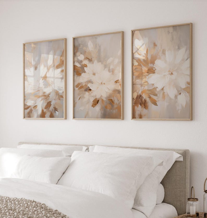 May include: Three framed abstract art prints with a beige background and white floral designs. The prints are arranged in a row on a white wall above a bed with white bedding.