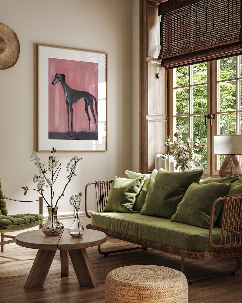 Puede incluir: Interior de sal&oacute;n con una obra de arte enmarcada de un galgo sobre un fondo rosa. La habitaci&oacute;n incluye un sof&aacute; verde con cojines, una mesa de centro de madera y un otom&aacute;n tejido. Una persiana de bamb&uacute; cubre la ventana.