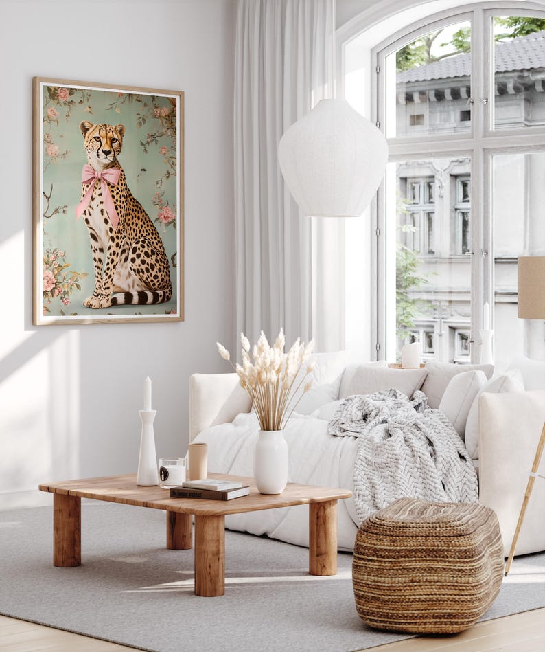 May include: A framed print of a cheetah wearing a pink bow tie, sitting on a light blue background with pink flowers. The cheetah is looking to the right. The print is hanging on a white wall in a living room setting.