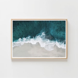May include: A framed aerial view of a beach with turquoise water and white waves crashing onto a sandy shore. The image is framed in a light-colored wooden frame, creating a serene and natural aesthetic.