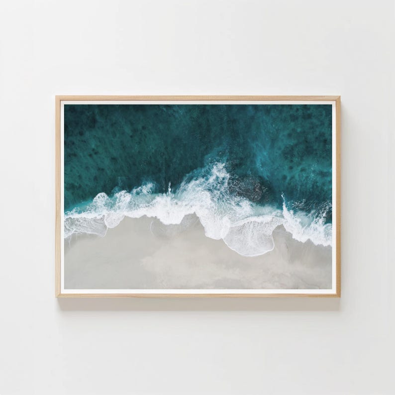 May include: A framed aerial view of a beach with turquoise water and white waves crashing onto a sandy shore. The image is framed in a light-colored wooden frame, creating a serene and natural aesthetic.