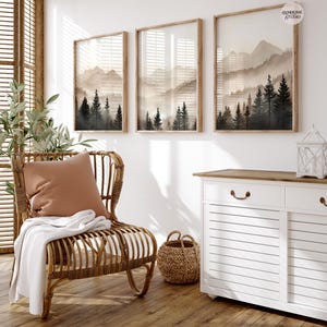 May include: Three vertical framed wall art prints featuring a misty mountain landscape with dark evergreen trees in the foreground and light beige peaks in the background. Each print is encased in a light wood frame and hung on a white wall.
