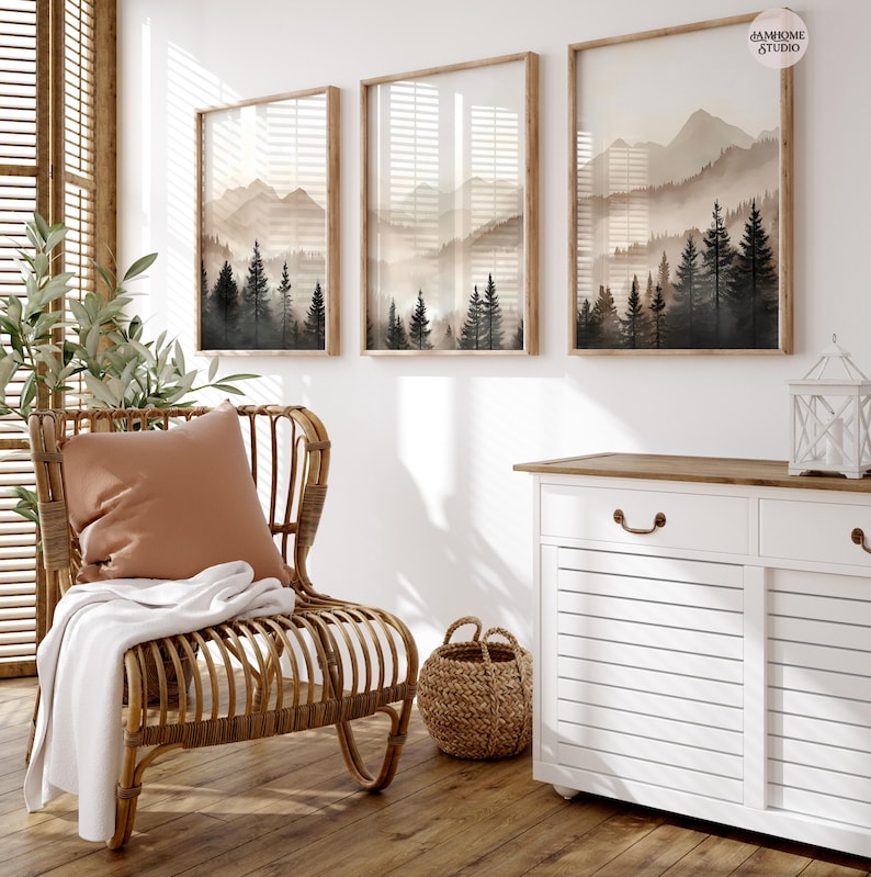 May include: Three vertical framed wall art prints featuring a misty mountain landscape with dark evergreen trees in the foreground and light beige peaks in the background. Each print is encased in a light wood frame and hung on a white wall.