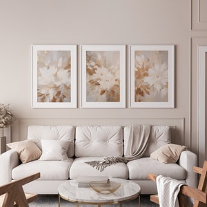May include: Three abstract art prints with a white background and brown and beige brushstrokes. The prints are framed in white and are hanging on a light beige wall above a white sofa.