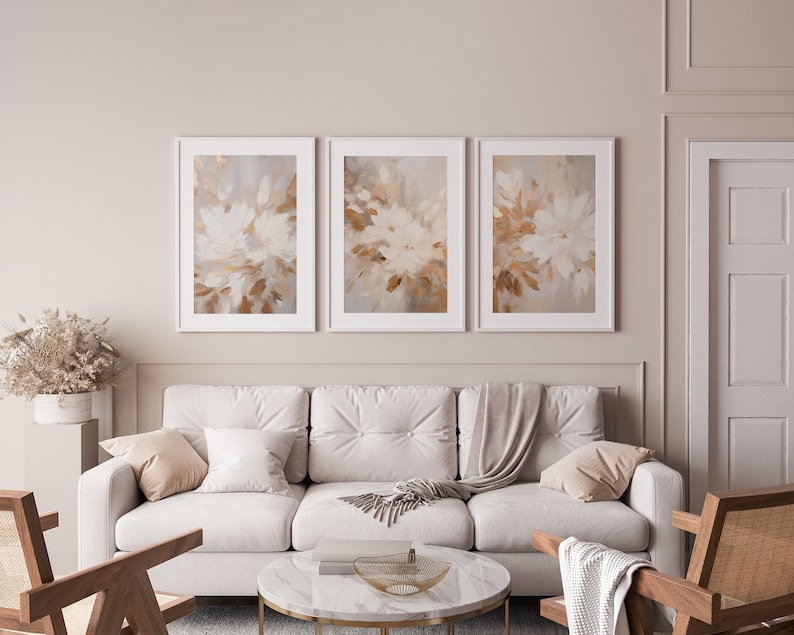 May include: Three abstract art prints with a white background and brown and beige brushstrokes. The prints are framed in white and are hanging on a light beige wall above a white sofa.