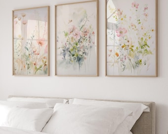 May include: Three framed floral art prints. Each print features a watercolor painting of delicate flowers in shades of pink, peach, and green. The frames are a light wood color, and the artwork is displayed above a bed with white bedding.