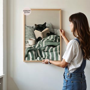 May include: A framed print depicts a black cat reading a book in bed, surrounded by green and white striped bedding and pillows. A white mug sits on the bed. The frame is light brown. The print is being held by a person.