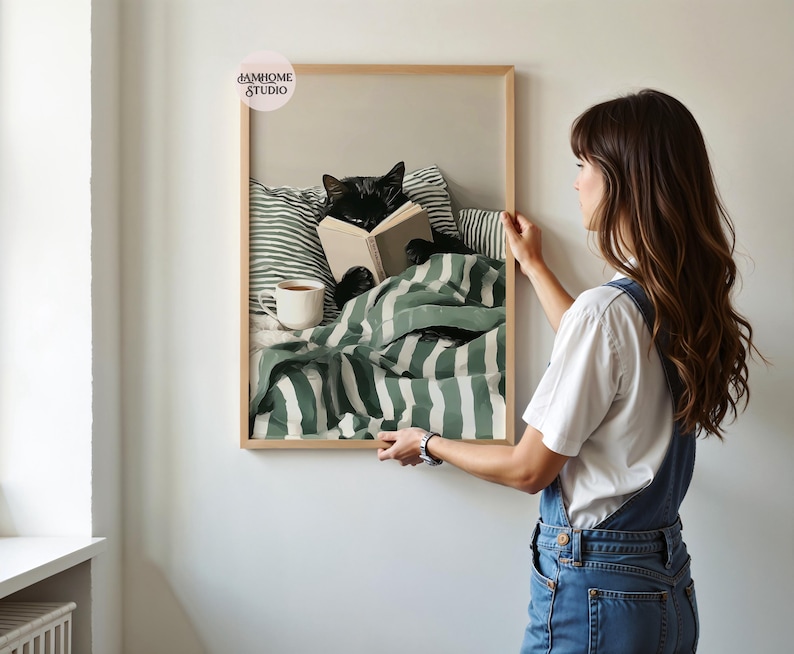 May include: A framed print depicts a black cat reading a book in bed, surrounded by green and white striped bedding and pillows. A white mug sits on the bed. The frame is light brown. The print is being held by a person.