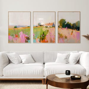 May include: A set of three rectangular wall art prints featuring impressionistic landscape paintings. The artwork displays fields of green grass, pink wildflowers, and a white house with a roof, rendered in soft, blended brushstrokes and colors.