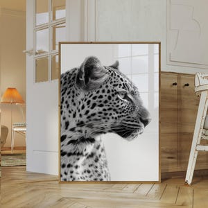 Può includere: Una stampa in bianco e nero incorniciata di un leopardo di profilo. L'opera d'arte presenta il mantello maculato e i dettagli del viso dell'animale. La cornice è in legno chiaro, aggiungendo un tocco di eleganza. La stampa è esposta in interni.