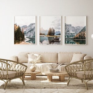 May include: Three framed landscape photographs of a mountain lake with snow-capped peaks and trees. The photos are hung on a white wall above a beige sofa with two wicker chairs and two coffee tables.