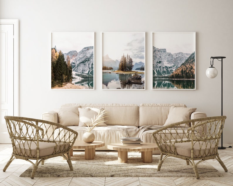 May include: Three framed landscape photographs of a mountain lake with snow-capped peaks and trees. The photos are hung on a white wall above a beige sofa with two wicker chairs and two coffee tables.