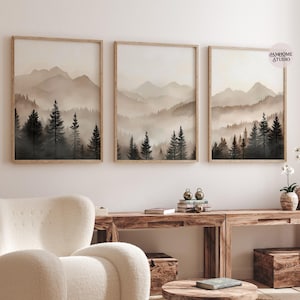 May include: A set of three framed wall art prints featuring a landscape of dark evergreen trees in the foreground and layered, misty mountains in the background. The color palette consists of muted beige, cream, and dark brown tones.