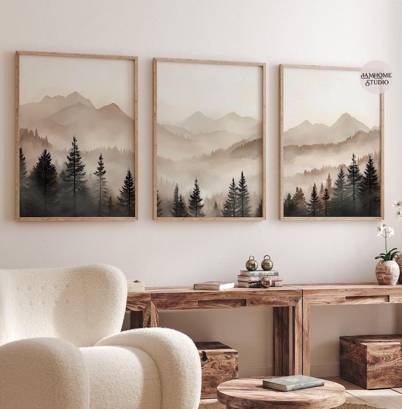 May include: A set of three framed wall art prints featuring a landscape of dark evergreen trees in the foreground and layered, misty mountains in the background. The color palette consists of muted beige, cream, and dark brown tones.
