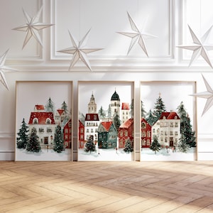 Könnte beinhalten: Drei gerahmte Kunstdrucke zeigen eine winterliche Dorflandschaft mit schneebedeckten Dächern und immergrünen Bäumen. Die Gebäude sind in Rot, Grün und Weiß gehalten. Sternförmige Ornamente hängen von der Decke.