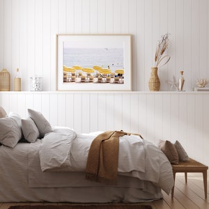 Puede incluir: Una impresi&oacute;n enmarcada de una escena de playa con sombrillas amarillas y tumbonas. La impresi&oacute;n cuelga sobre una cama con ropa de cama blanca y una manta marr&oacute;n. Objetos decorativos en una estanter&iacute;a.
