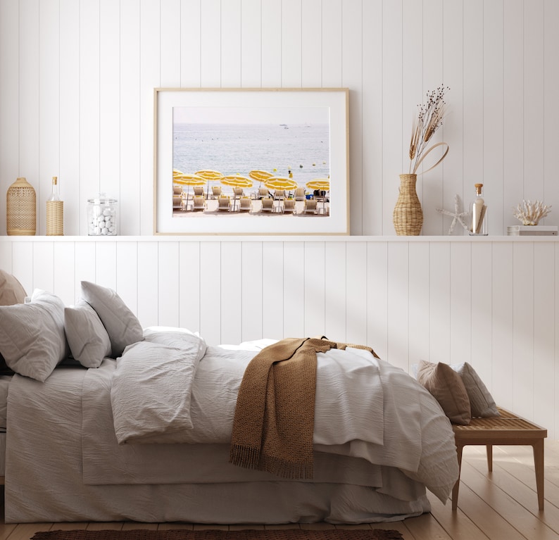Puede incluir: Una impresi&oacute;n enmarcada de una escena de playa con sombrillas amarillas y tumbonas. La impresi&oacute;n cuelga sobre una cama con ropa de cama blanca y una manta marr&oacute;n. Objetos decorativos en una estanter&iacute;a.
