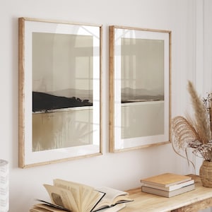 Puede incluir: Dos impresiones artísticas enmarcadas de paisajes en tonos neutros. Cada impresión presenta un diseño minimalista con una colina oscura y texturizada, una masa de agua clara y un cielo apagado. Los marcos son de madera clara.