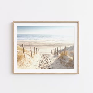 Könnte beinhalten: Ein gerahmter Druck einer Strandszene. Ein sandiger Pfad führt zum Meer, gesäumt von Holzpfosten und Dünen mit hohem Gras. Der Himmel ist hellblau und die Gesamtpalette ist neutral mit warmen Tönen.