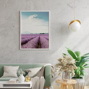 May include: A framed photograph of a lavender field with a blue sky and white clouds. The photo is hanging on a white wall above a green sofa with white pillows and a throw blanket. There is a small table with a vase of white flowers and a gold decorative object.