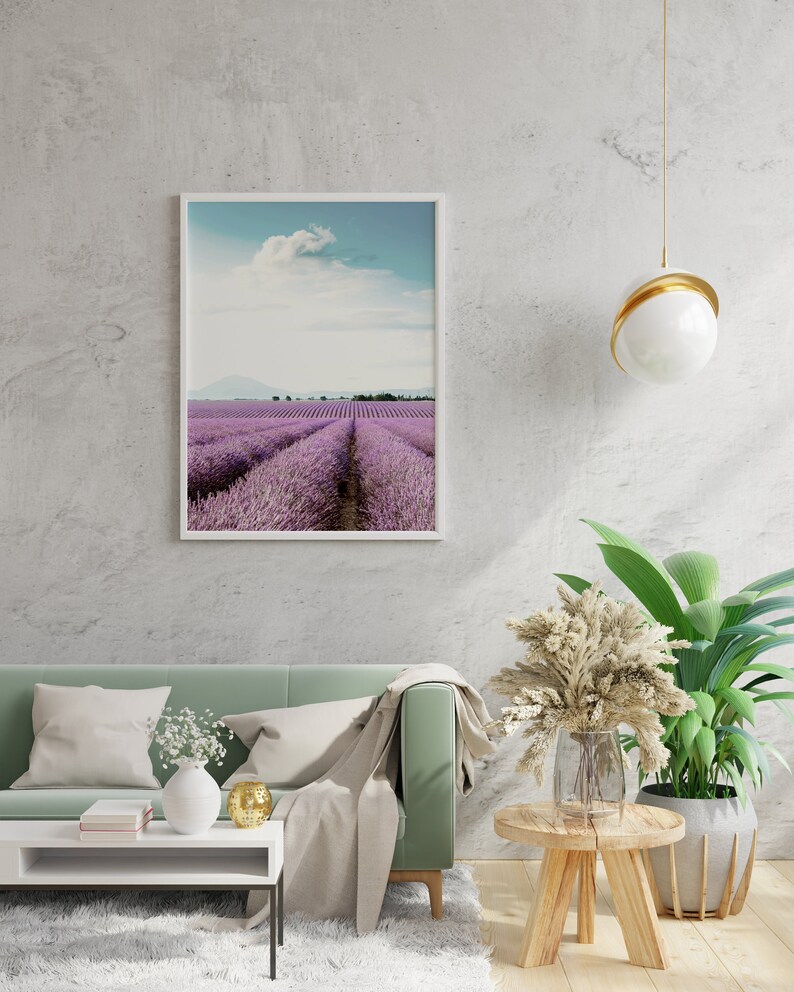 May include: A framed photograph of a lavender field with a blue sky and white clouds. The photo is hanging on a white wall above a green sofa with white pillows and a throw blanket. There is a small table with a vase of white flowers and a gold decorative object.