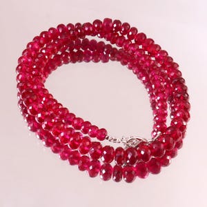 May include: A multi-strand necklace composed of small, faceted, deep red beads. The beads are uniform in size and shape, creating a reflective, textured appearance. The necklace is secured with a silver-toned metal lobster claw clasp.