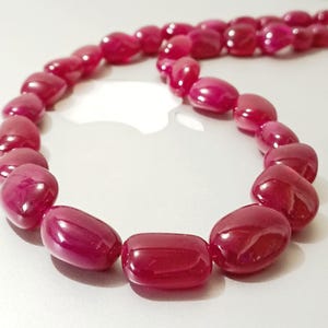 May include: A necklace composed of polished, deep red gemstones. The stones vary in shape, with some being rounded and others more rectangular. The necklace is displayed against a light background, highlighting the rich color of the stones.