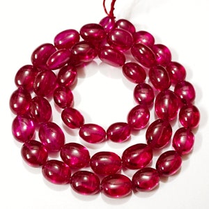 May include: A strand of polished, oval-shaped beads in a deep, translucent magenta-red color. The beads are strung together on a thin red cord, arranged in a circular pattern with a smooth, glossy finish that reflects light across their surfaces.