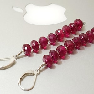 May include: A pair of dangle earrings featuring faceted, deep red gemstones strung on a silver wire. The earrings have a lever-back closure. The gemstones are a vibrant, rich color, and the earrings are displayed on a light gray surface.