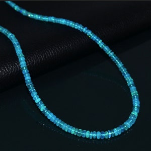 Puede incluir: Un collar hecho de peque&ntilde;as cuentas redondas de &oacute;palo azul claro. El collar est&aacute; sobre una superficie negra.