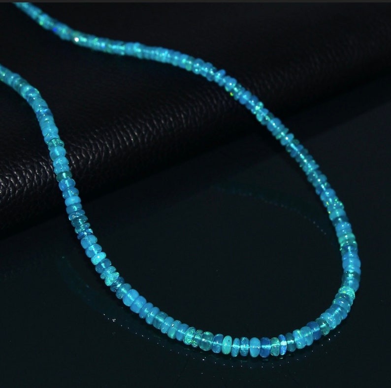 Puede incluir: Un collar hecho de peque&ntilde;as cuentas redondas de &oacute;palo azul claro. El collar est&aacute; sobre una superficie negra.