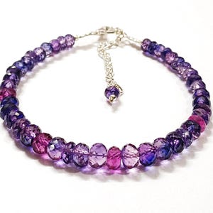 Op de afbeelding: Een kralenarmband met een verloop van paarse en roze edelstenen. De armband heeft een zilverkleurige ketting en sluiting. De edelstenen zijn facetgeslepen en reflecteren licht. Sieraad voor de pols.