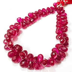 May include: A strand of faceted, teardrop-shaped ruby beads. The beads are a deep, rich red color and are strung together. A red tassel is attached at the top. The beads are displayed against a white background.