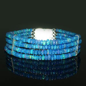 Op de afbeelding: Een armband met meerdere strengen bestaande uit vier rijen kleine, ronde, blauwe opaal kralen. De kralen vertonen iriserende flitsen van groen en roze. De strengen worden bij elkaar gehouden door twee verticale zilverkleurige metalen staven en een sluiting.