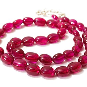 May include: A strand of oval-shaped ruby red beads strung on a silver chain. The beads are all the same size and shape, and they are arranged in a symmetrical pattern.