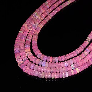 May include: A strand of pink opal beads, with a slight iridescent shimmer, is shown in a close-up image. The beads are round and smooth, and they are arranged in a loose, flowing pattern.