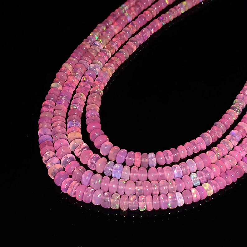 May include: A strand of pink opal beads, with a slight iridescent shimmer, is shown in a close-up image. The beads are round and smooth, and they are arranged in a loose, flowing pattern.
