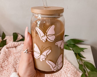 Puede incluir: Un tarro de vidrio con tapa de bambú y una pajita de vidrio. El tarro está lleno de un líquido marrón y tiene un diseño de mariposa rosa y dorada.