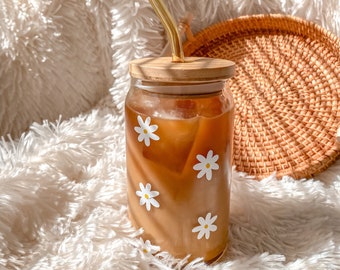 Puede incluir: Un tarro de vidrio con tapa de madera y una pajita lleno de café helado. El tarro tiene un patrón de margaritas y está sentado sobre una superficie blanca y esponjosa.