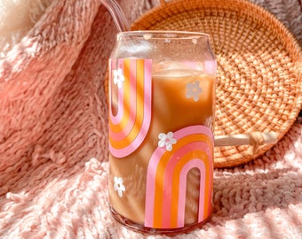 Puede incluir: Un vaso con un diseño de arcoíris y flores lleno de una bebida marrón claro. Una pajita de vidrio está en el vaso. El vaso está sobre una superficie rosa y peluda.