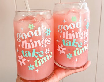 Puede incluir: Dos vasos de vidrio transparente llenos de líquido rosa y cubitos de hielo. Los vasos tienen un fondo blanco con el texto "good things take time" en letras rosa, azul y verde. Cada vaso tiene una pajita de vidrio transparente.