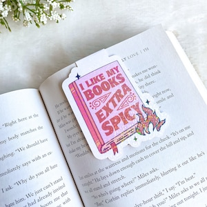 May include: A pink and white bookmark with glitter accents. The bookmark features a book with the text "I Like My Books Extra Spicy" and a flame design. The bookmark is on a page of a book with text.