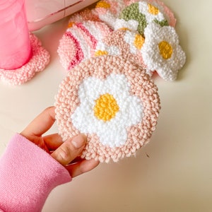 Könnte beinhalten: Ein Set handgemachter Untersetzer in Form von Gänseblümchen in den Farben Rosa, Weiß und Gelb. Die Untersetzer sind aus einem flauschigen Garn gefertigt und haben eine erhabene Mitte.