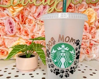 May include: A white reusable cup with a black and green Starbucks logo and the text "Dog Mom" in gold lettering. The cup is surrounded by black paw prints.