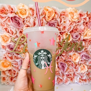 May include: A clear plastic reusable cup with a pink and white star design and a pink Pisces zodiac symbol. The cup is filled with iced coffee and has a pink straw.